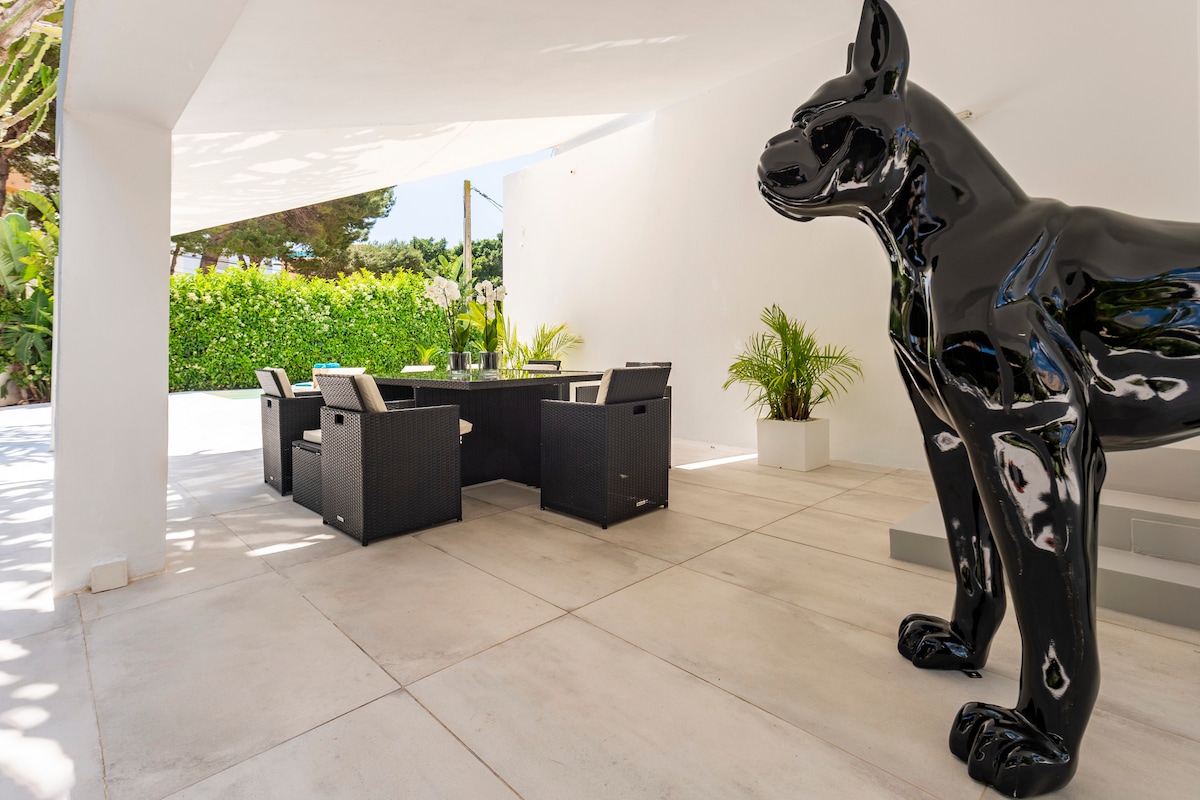 An outdoor dining area is surrounded by lush greenery, featuring a large table with black wicker chairs. A sleek black dog sculpture enhances the modern aesthetic. The space is shaded, creating a comfortable atmosphere for relaxation or meals.