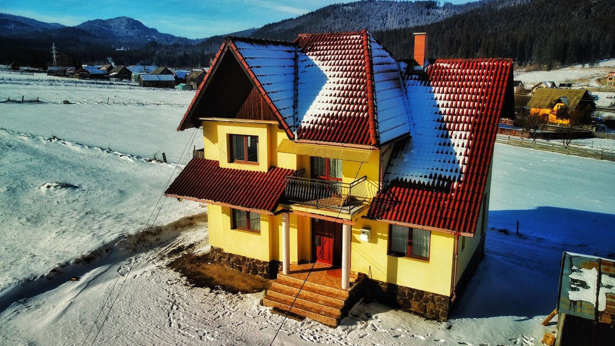 A spacious two-story house with a red-tiled roof is nestled in a snowy landscape. The exterior features yellow walls and a stone foundation. Surrounding fields are covered in fresh snow, with mountains visible in the background under a clear blue sky.