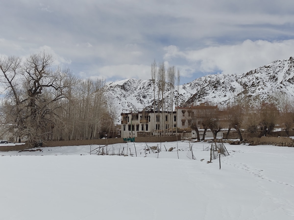 A historic building stands amidst a snowy landscape, surrounded by barren trees and distant mountains. The structure features a faded exterior with several windows, and a fence encloses a partially visible area in front, adding to the tranquil winter scene.