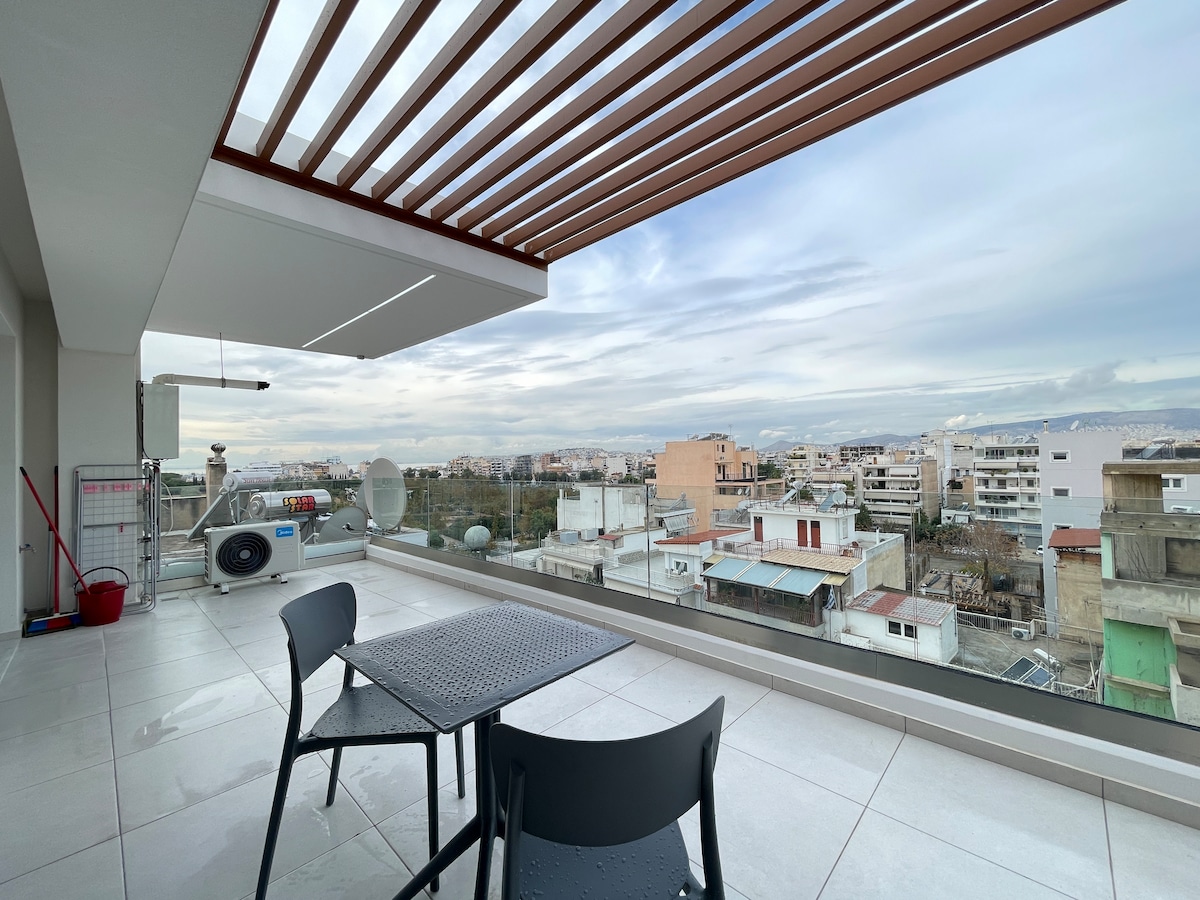 An outdoor terrace is shown with a table and two chairs under a slatted awning. The space offers a panoramic view of the surrounding neighborhood, featuring various building rooftops and a distant Athens skyline. The flooring consists of large, light-colored tiles.