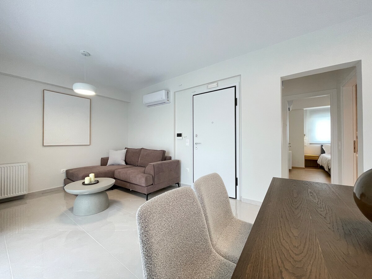 A modern living area is presented with a comfortable sectional sofa facing a round coffee table. Light from a ceiling fixture illuminates the space, while a door leads to an adjacent room. A dining table with two chairs is positioned nearby, creating a functional layout.