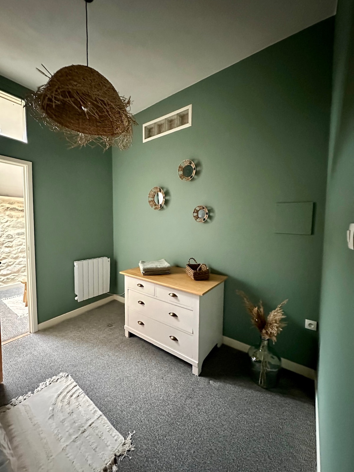 A welcoming entryway features a white dresser with five drawers and a wooden top. Three circular mirrors are mounted on an olive green wall, reflecting light. A neutral rug lies on the gray carpeted floor, while a glass vase with dried plants adds a touch of nature.