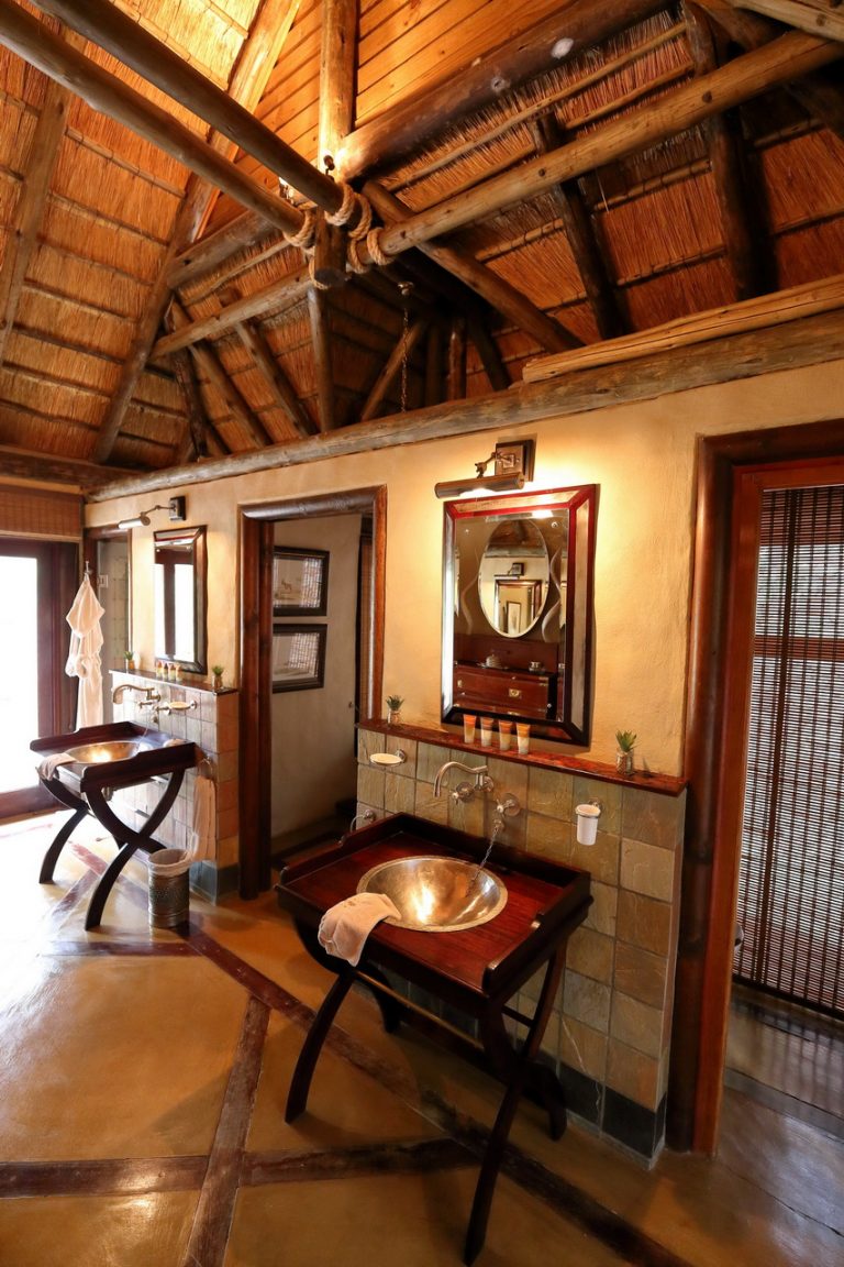 The bathroom features a rustic design with high thatched ceilings and wooden accents. A modern sink with a polished silver bowl is positioned on a wooden console. Mirrors are mounted on the walls, complemented by subtle lighting that enhances the natural materials used throughout the space.