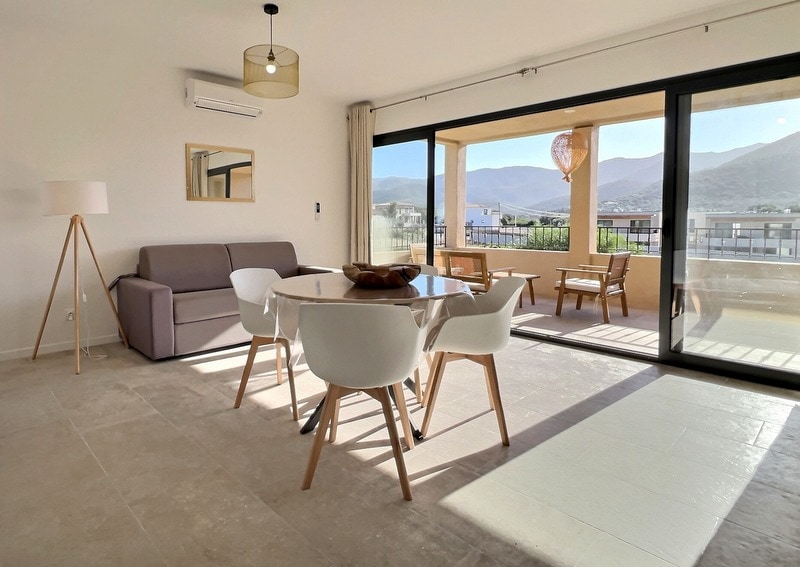 A bright interior space features a dining area with a round table surrounded by four white chairs, complemented by a modern sofa. Large glass doors open to a balcony showcasing scenic mountain views, while natural light fills the room, enhancing the open and airy atmosphere.