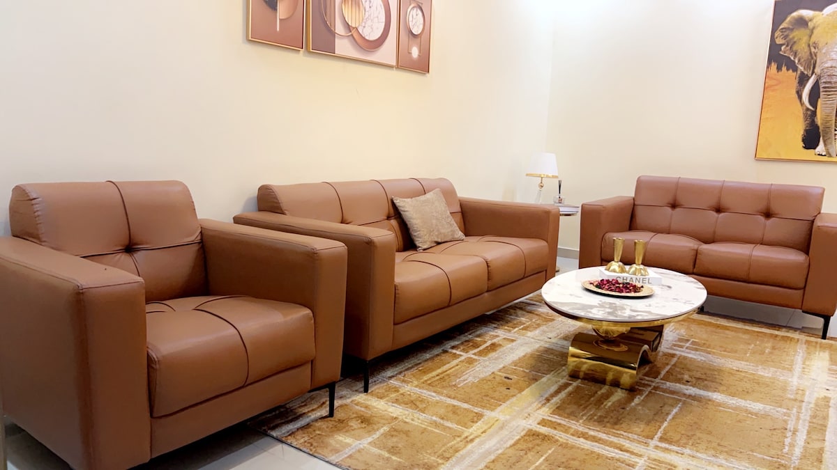 A modern living area is furnished with two brown leather sofas and an upholstered armchair, complemented by a round marble coffee table. An area rug adds warmth underfoot, and elegant wall art depicting an elephant enhances the ambiance.