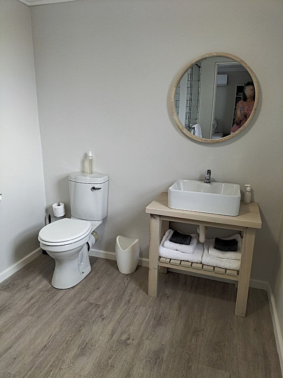 A clean bathroom is presented, featuring a modern toilet and a wooden vanity with a white basin. Beneath the basin, neatly stacked towels are displayed. A round mirror hangs above the sink, enhancing the spacious feel of the area. Light wooden flooring completes the design.