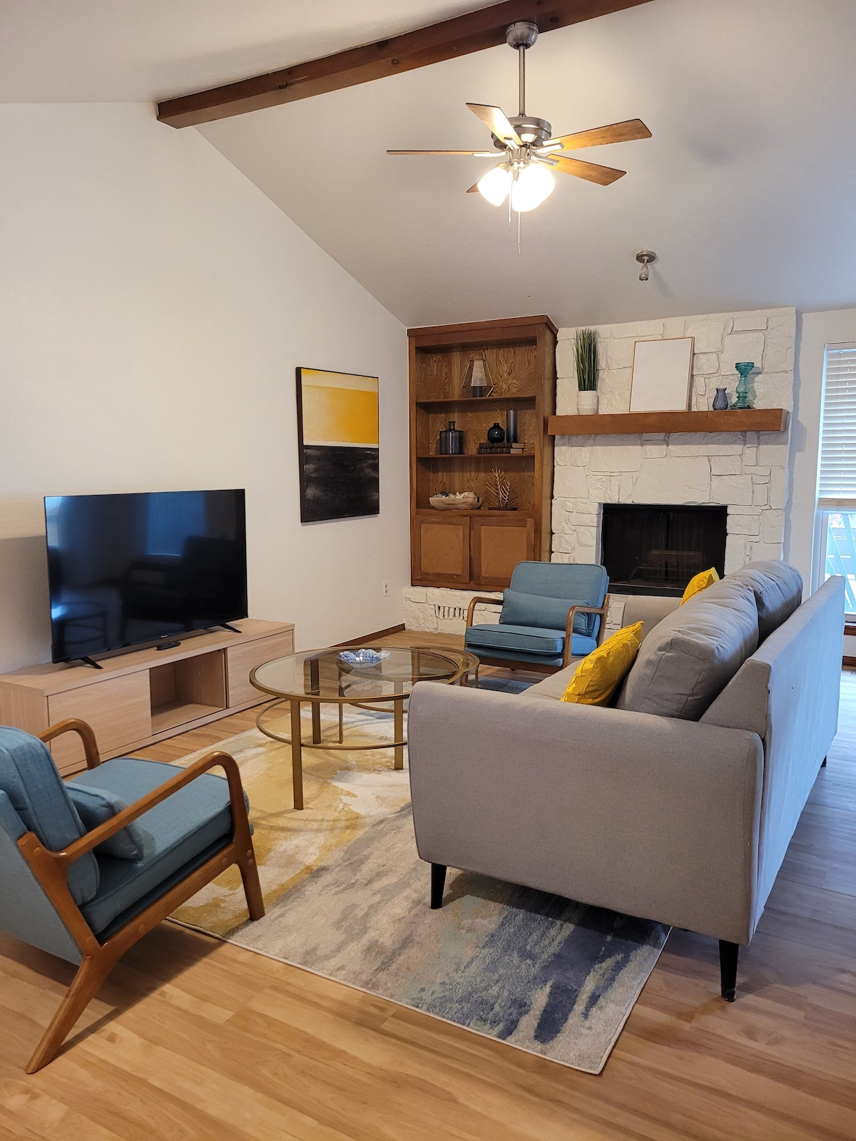 The living area features a light-colored sofa and two blue armchairs arranged around a glass coffee table. A television stands on a wooden console against one wall, while a stone fireplace adds texture and warmth to the space. Natural light filters through nearby windows.