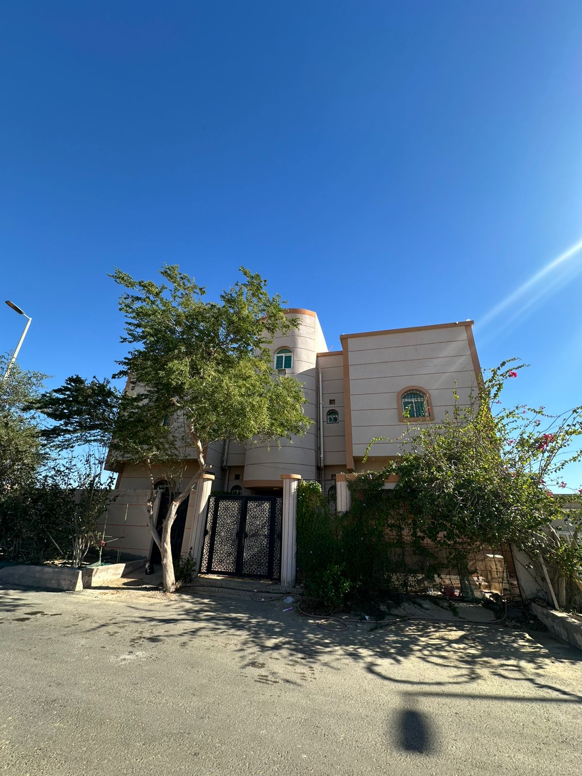 A charming two-story building is surrounded by greenery, with a mix of trees and shrubs. The entrance features a decorative gate that opens to a pathway leading to the front door. Clear blue skies above provide a bright backdrop.