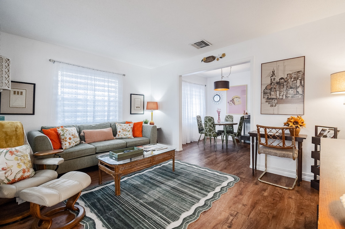 A cozy living room features a comfortable sofa adorned with colorful throw pillows, paired with a lounge chair. Natural light filters through sheer curtains, illuminating the wooden floor. A dining area with patterned chairs is visible through the archway, enhancing the inviting atmosphere.