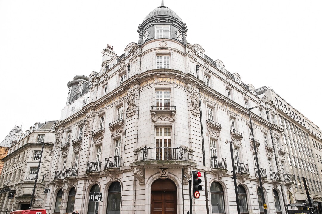 A historic building in the city center features intricate architectural detailing, including decorative moldings and multiple balconettes. The grand entrance is framed by ornate stonework, and large windows can be seen throughout the façade, allowing for ample natural light.
