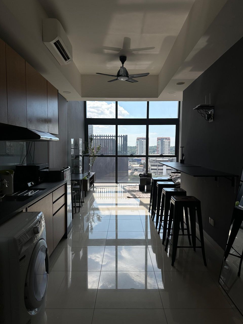 A modern kitchen area is visible, featuring sleek wooden cabinets and appliances including a washing machine. A row of tall stools lines a bar table by large windows that allow natural light to fill the space, showcasing a view of the city below.