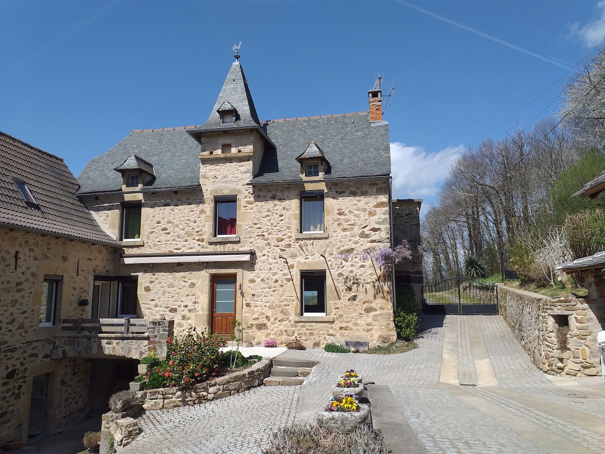 The exterior of a charming stone house with a pointed roof is highlighted. Surrounded by a well-maintained garden, colorful flowers line the pathway leading to the entrance. A gravel driveway extends toward a gated area in the background, framed by trees under a clear blue sky.