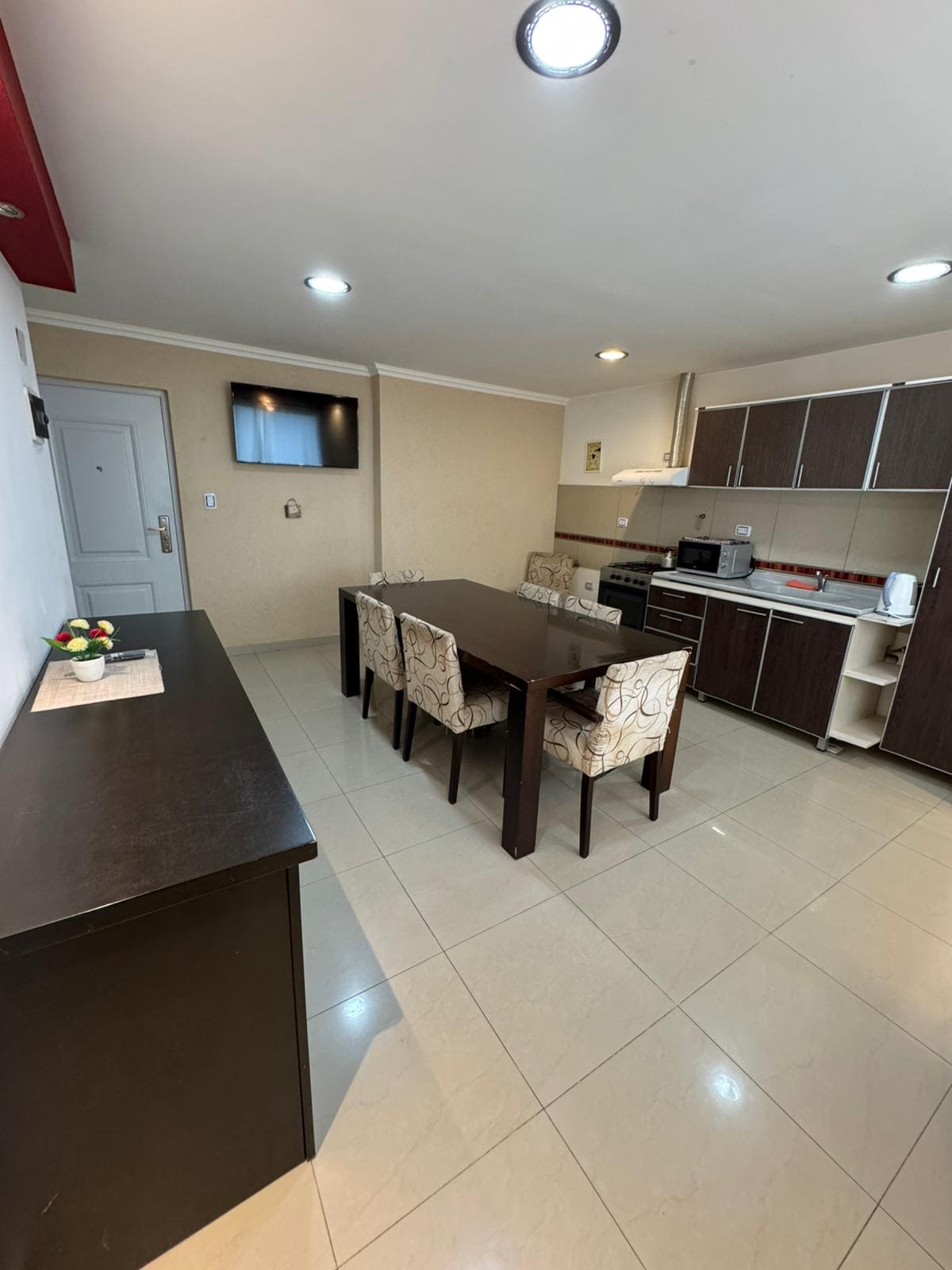 A modern kitchen and dining area is displayed, featuring a long wooden table surrounded by four patterned chairs. The kitchen cabinetry is dark brown, and a stainless steel stove is visible. Bright overhead lights illuminate the space, enhancing its practicality.