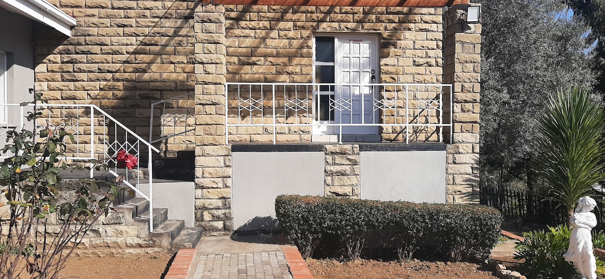 A stone exterior of the guesthouse is highlighted, featuring a white door framed by a railing and accessible steps. The neatly landscaped area includes low shrubs and a pathway made of brick, contributing to the welcoming entrance.