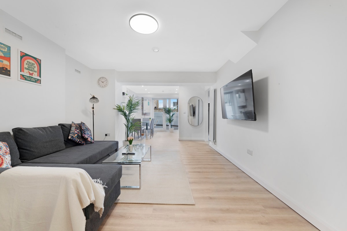A modern living area is showcased with a dark sectional sofa adorned with patterned cushions, facing a wall-mounted television. A glass coffee table is positioned on a light area rug, while a plant adds a touch of greenery. The space is filled with natural light.