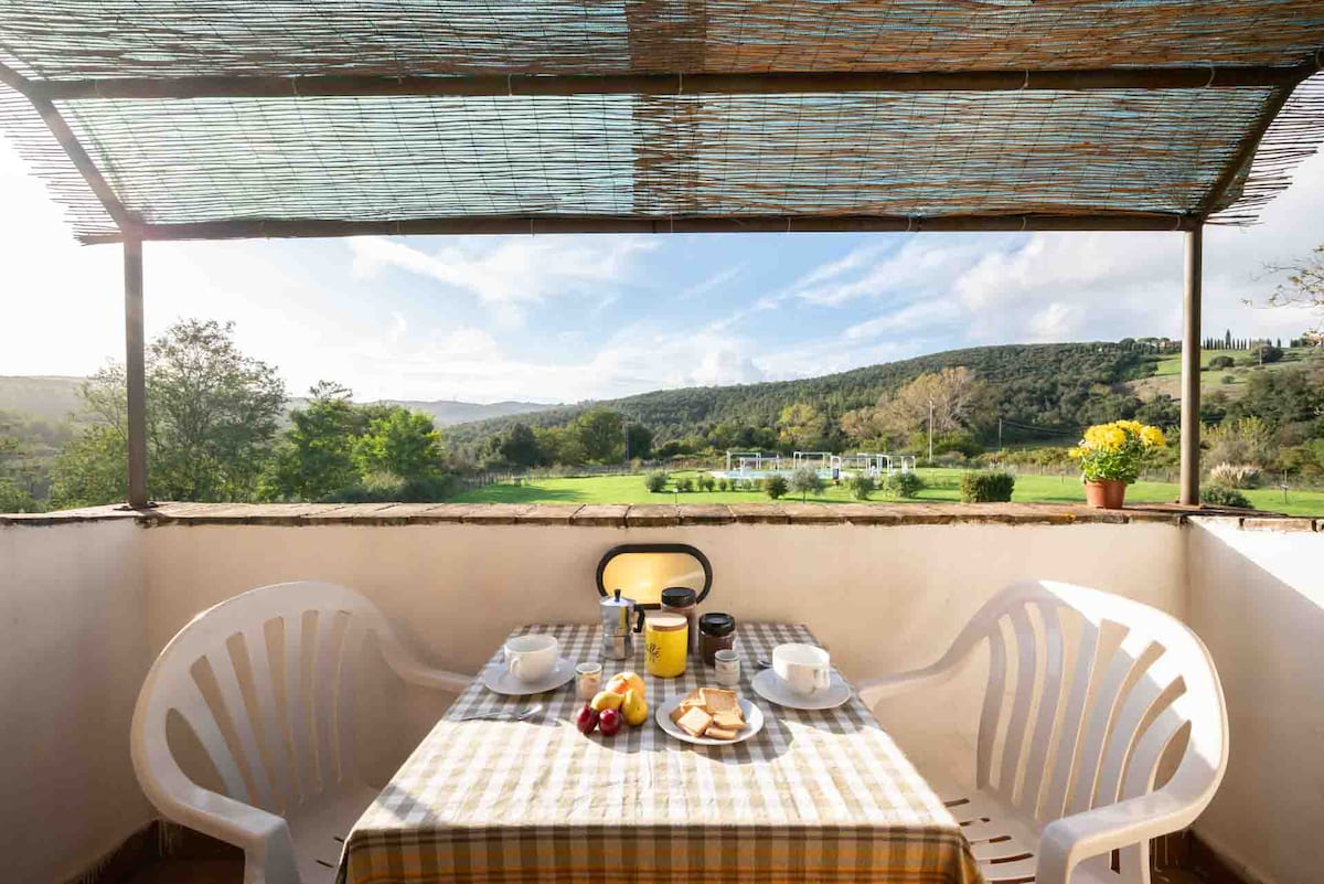 A private balcony features a table set for breakfast, adorned with various pastries, fruit, and drinks. The space is shaded by a reed canopy, offering a view of lush green hills and gardens in the distance, providing a peaceful outdoor dining experience.