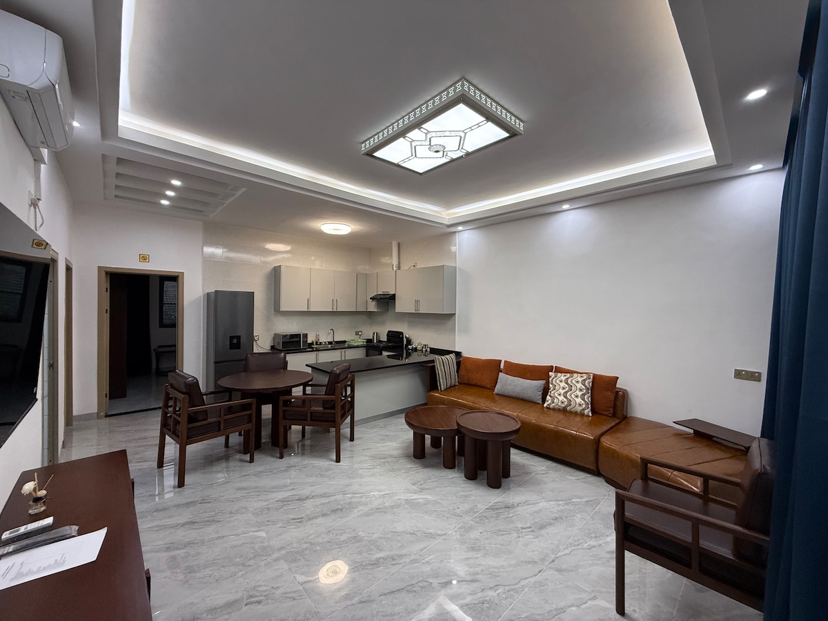 A modern living area features a brown leather sectional couch paired with wooden chairs and tables. Light-colored cabinetry complements the kitchen area, while a sleek ceiling with indirect lighting adds a contemporary touch. Walls are painted in a neutral shade, creating a spacious feel.