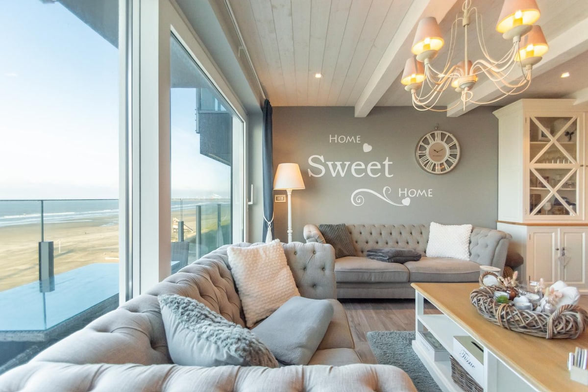 The living area features large windows offering views of the sea, complemented by a cozy seating arrangement. A clock and decorative wall art enhance the space's character. A warm light fixture hangs from the ceiling, creating a welcoming atmosphere throughout the room.