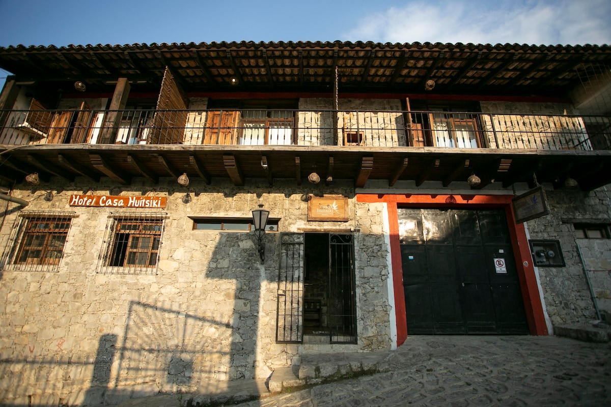 A rustic hotel exterior features a stone wall and wooden balcony. The entrance is framed by large wooden doors, with a lantern providing illumination. The building's design reflects traditional architecture, with small windows above and decorative metalwork enhancing the facade.
