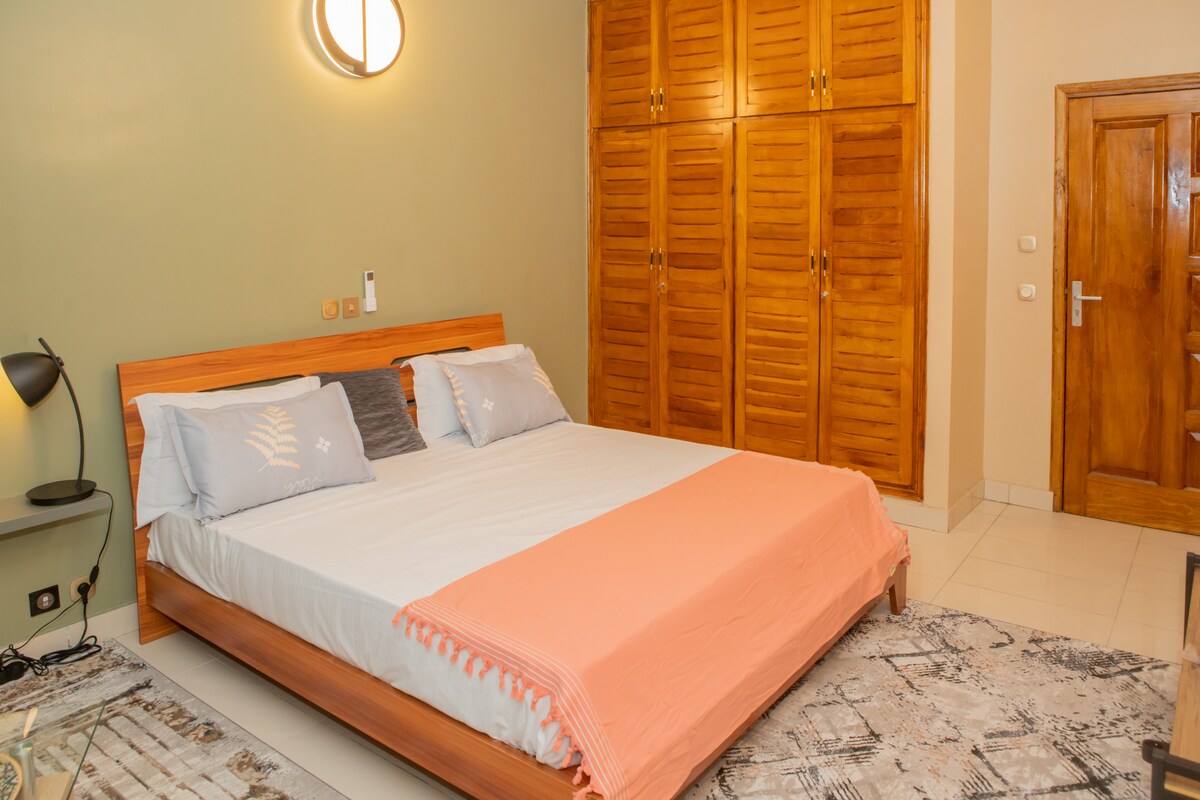 The spacious sleeping area features a wooden bed framed by soft bedding in neutral colors. A large wooden wardrobe occupying a wall provides ample storage. Warm lighting is offered by a circular fixture above, enhancing the relaxing atmosphere. A textured rug adds a cozy touch to the floor.