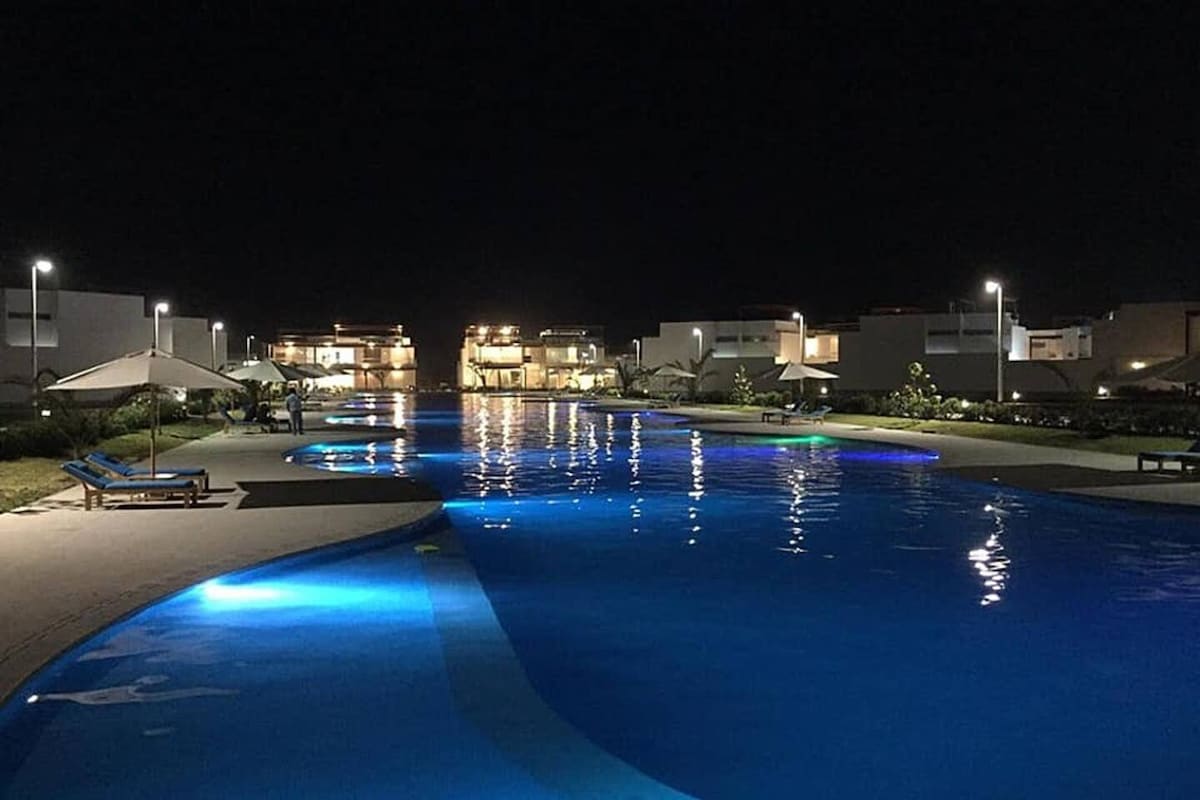 The nighttime view reveals a spacious swimming pool, illuminated with soft blue lights. Surrounding areas are landscaped with green grass and palm trees, while modern structures are visible in the distance. The tranquil atmosphere is enhanced by the reflection of lights on the water's surface.