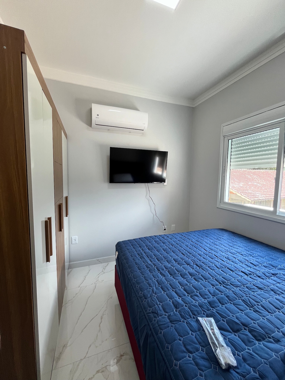 A serene bedroom features a spacious bed with a blue quilted cover and a wardrobe with wooden accents. A wall-mounted television is positioned opposite the bed, while natural light enters through a nearby window covered by a light curtain. An air conditioning unit is installed above.