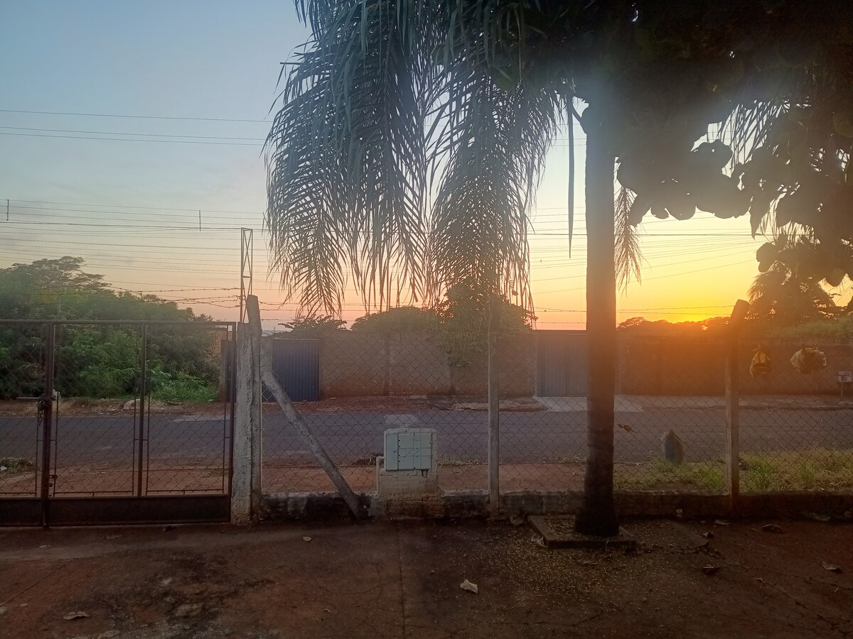 The image captures the exterior view of a fenced property at sunset. Soft hues of orange and pink in the sky highlight the silhouette of trees and a distant road. A palm tree and a clear pathway lead towards a gated entrance, creating a calm outdoor atmosphere.