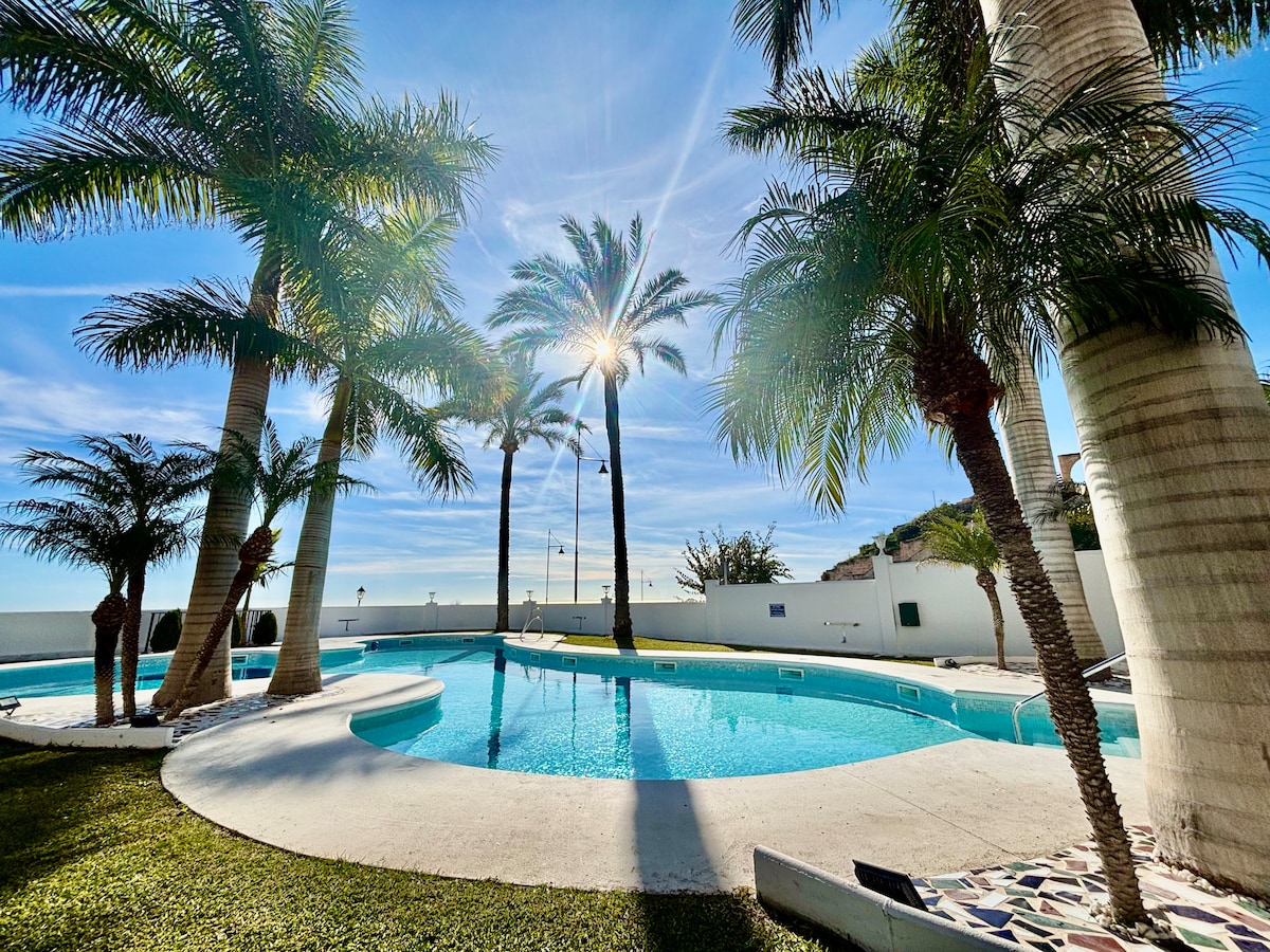 A well-maintained communal pool is surrounded by tall palm trees, casting soft shadows on the pristine water. The tranquil setting is bathed in sunlight, with the sky displaying a serene blue, creating a relaxing atmosphere ideal for leisure.