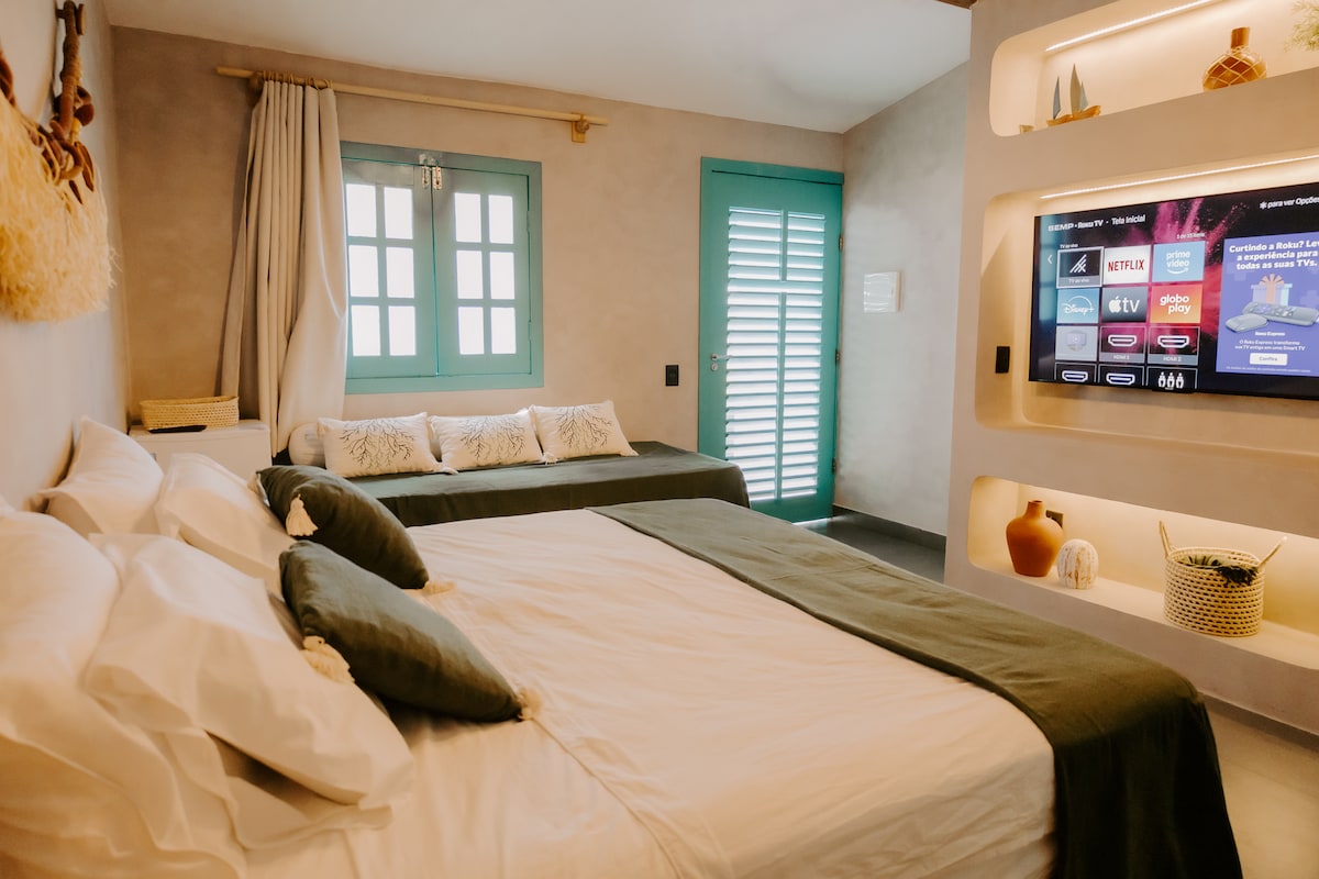 A stylish bedroom features a spacious bed adorned with soft linen and accent pillows. A single bed is positioned beside it. Natural light enters through a window, complemented by a vibrant turquoise door. A media console with a flat-screen TV is installed on the wall.