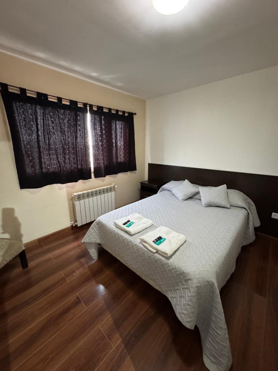 A well-appointed bedroom features a spacious bed with a light grey coverlet and decorative pillows. Two neatly folded towels are placed at the foot of the bed. Natural light enters through a window, which is dressed with sheer dark curtains, while a radiator adds comfort.