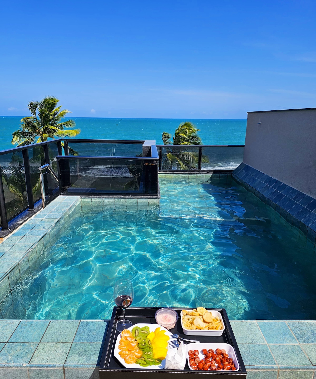 An inviting private rooftop pool appears, surrounded by clear blue waters and bright skies. A tray with a variety of snacks and drinks is positioned on the edge of the pool, enhancing the setting for relaxation or enjoyment.