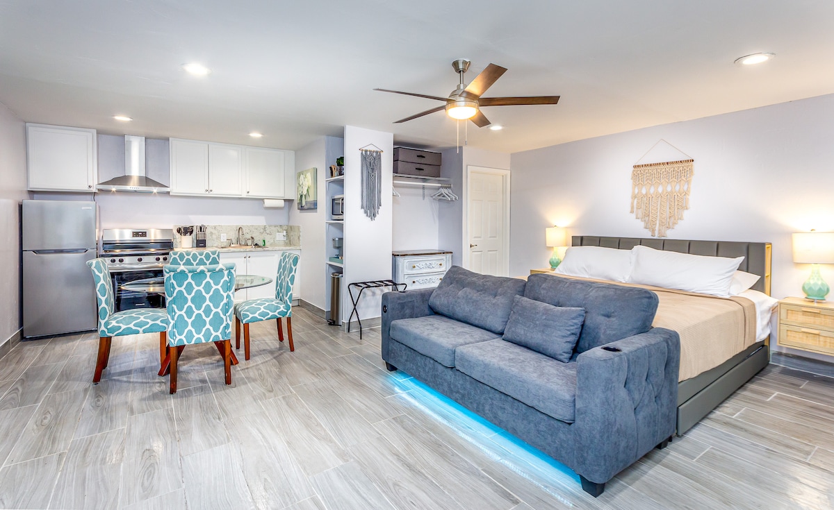 An open-concept studio features a spacious bed with light bedding and a cozy sofa in front. A dining area with two patterned chairs is positioned adjacent to the fully equipped kitchen. Natural light fills the room, highlighting the modern design and neutral color palette.