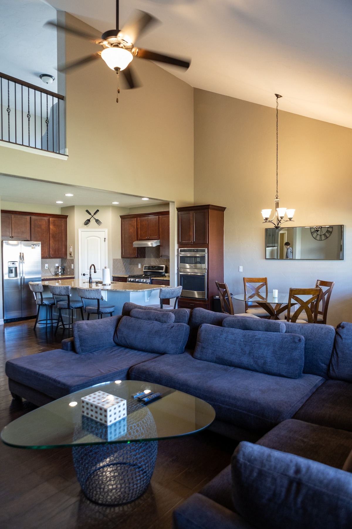 An open living area features a dark sectional sofa set around a glass coffee table. A modern kitchen with stainless steel appliances and a dining table for six is visible in the background. Ceiling fans and light fixtures add illumination to the space.