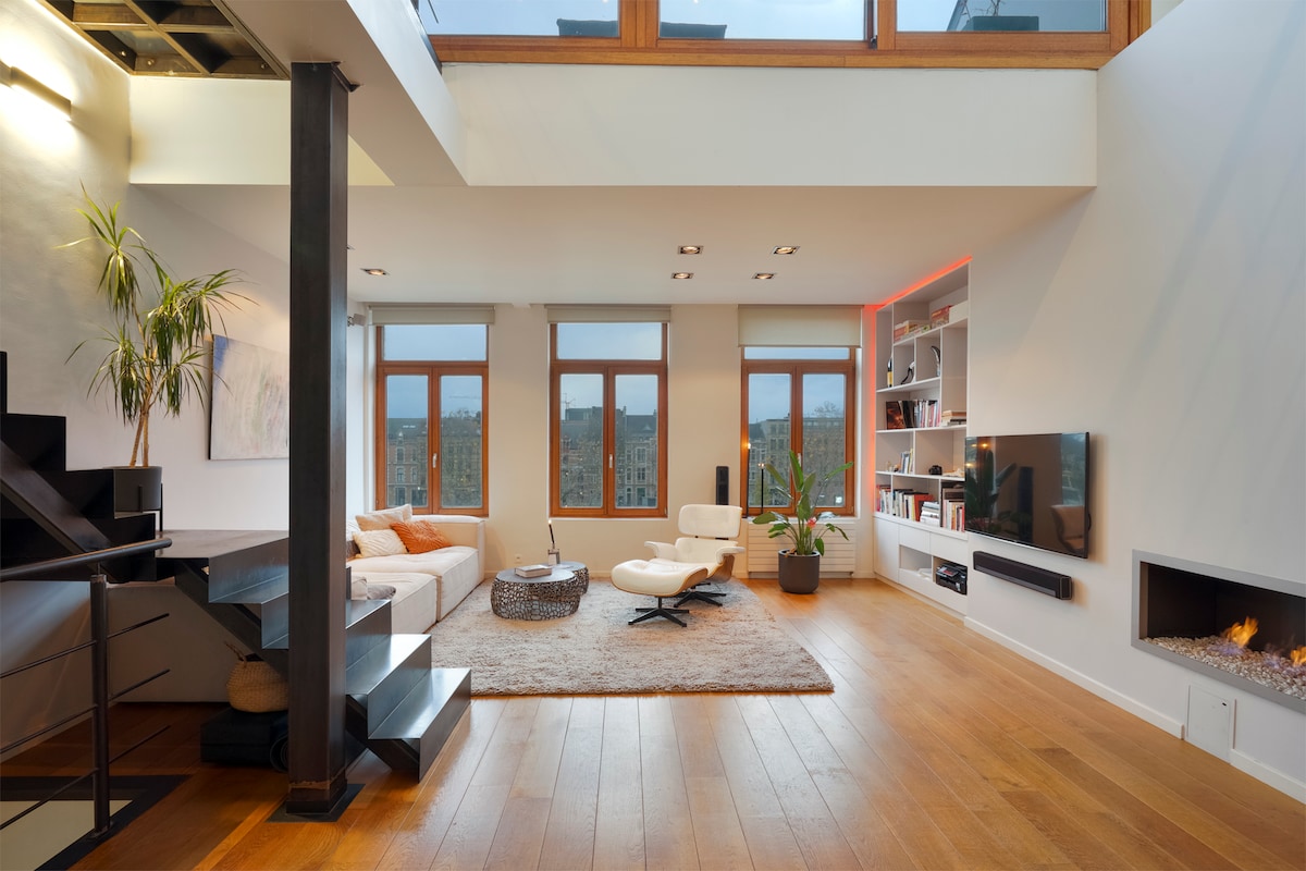 The spacious living area is highlighted by large windows that provide ample natural light and scenic views. A plush rug anchors the seating area, which includes a modern chair and a sectional sofa. A sleek television and a decorative fireplace are featured on the wall, surrounded by built-in shelves.