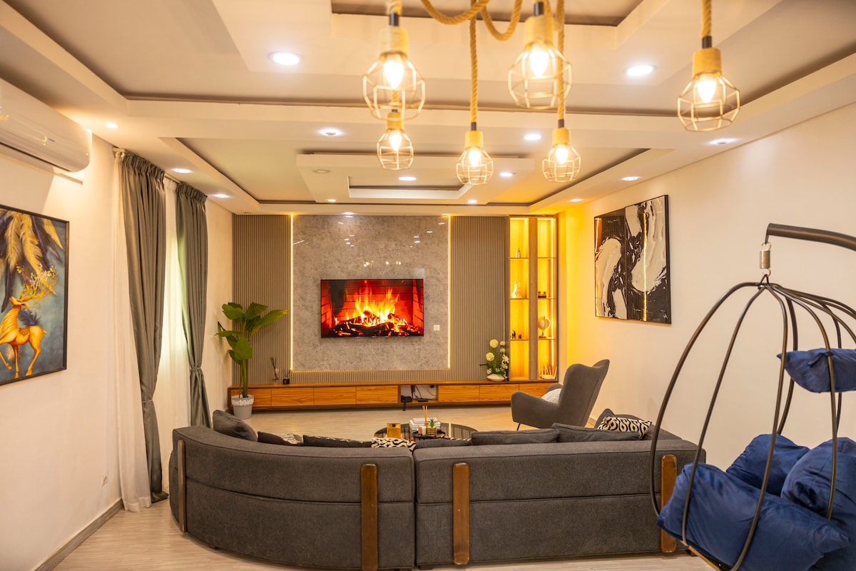 A spacious living area is furnished with a large, curved sofa and a hanging chair. A modern television is mounted on a feature wall, complemented by decorative lighting. Artwork adorns the walls, and soft curtains frame the windows, allowing natural light to fill the space.