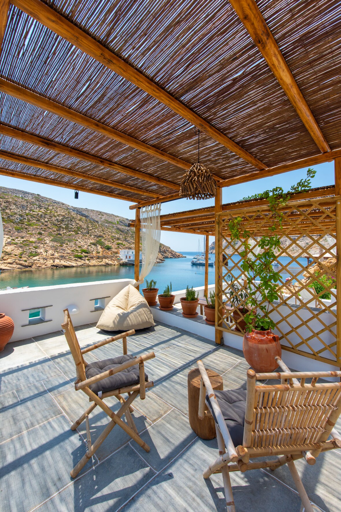 An outdoor terrace is presented, featuring a bamboo pergola that provides shade. Two foldable chairs and a bean bag are arranged on the tiled floor, surrounded by potted plants. A serene view of the coastal landscape and boats is visible in the background.