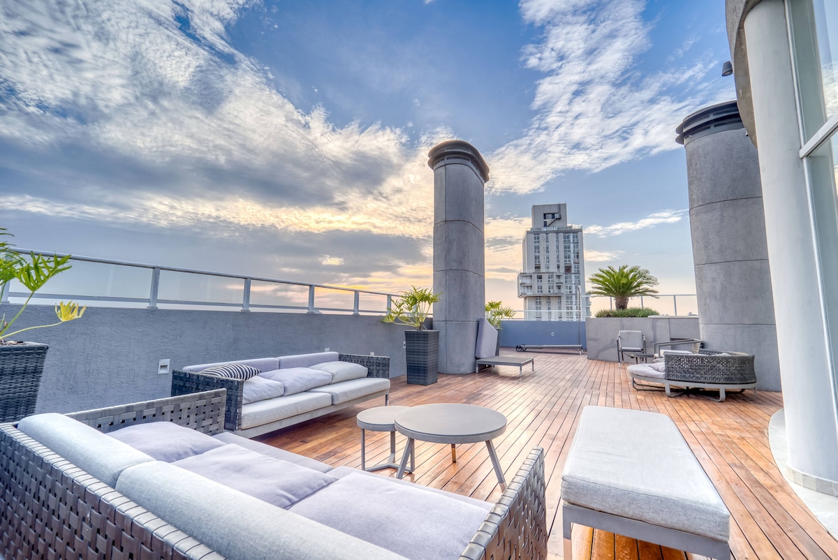 A rooftop terrace features stylish seating arrangements, including a spacious sectional sofa and a coffee table. The wooden deck is surrounded by planters, and views of the sky and the city can be enjoyed from this elevated space.
