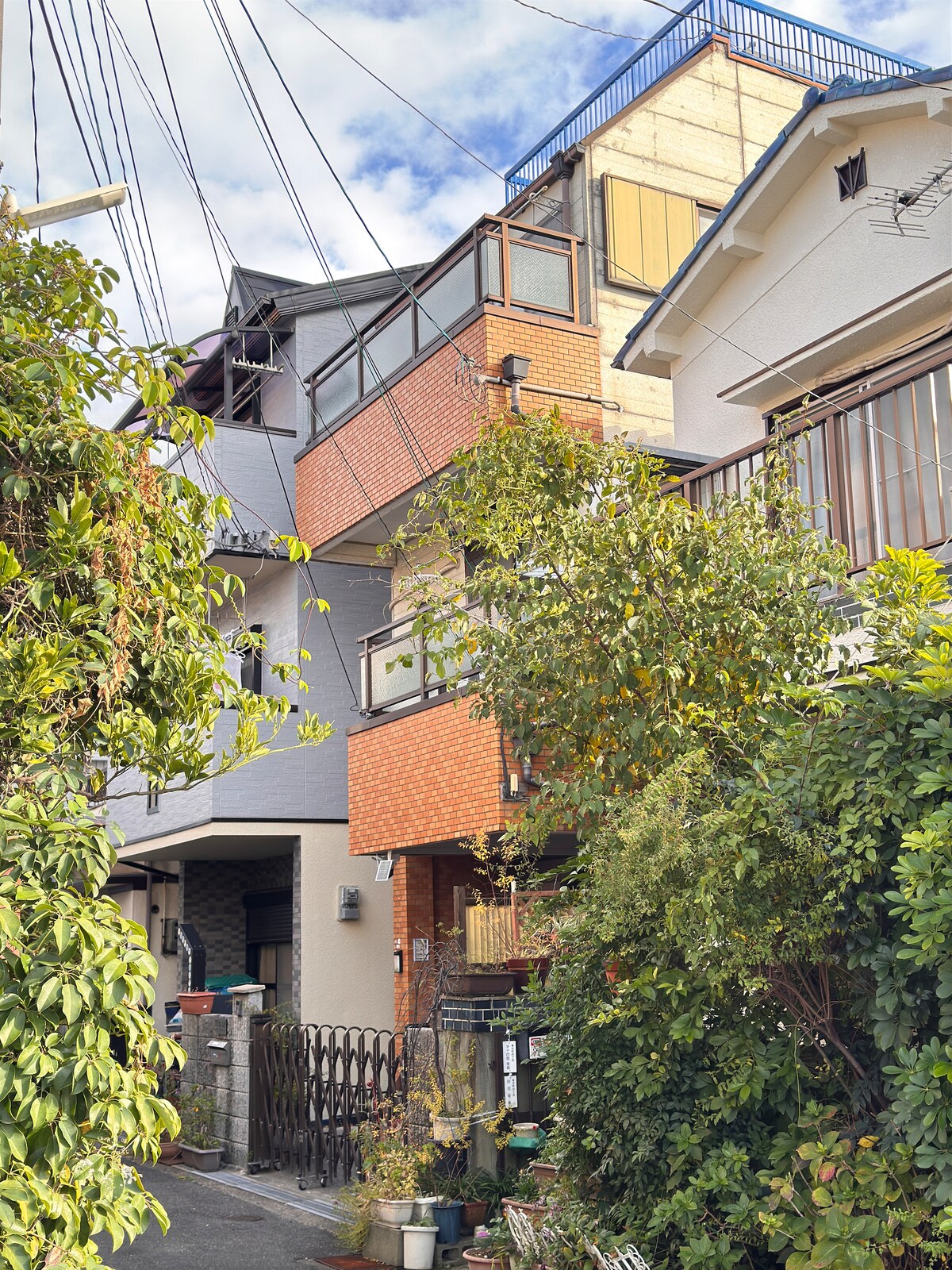 A multi-story residential building showcases a blend of architectural styles, with brick and plaster facades. Green foliage lines the pathway, enhancing the inviting atmosphere of the neighborhood. A gated entrance provides access, with power lines overhead and a clear sky in the background.