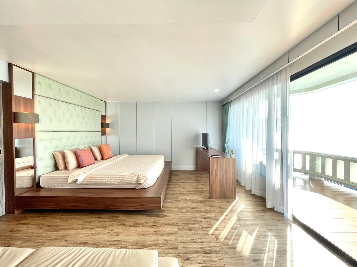 A spacious room features a large bed with light-colored linens and accent pillows in warm tones. A wooden entertainment unit is positioned nearby, and floor-to-ceiling windows create a bright atmosphere, offering views of a balcony area framed by sheer curtains.