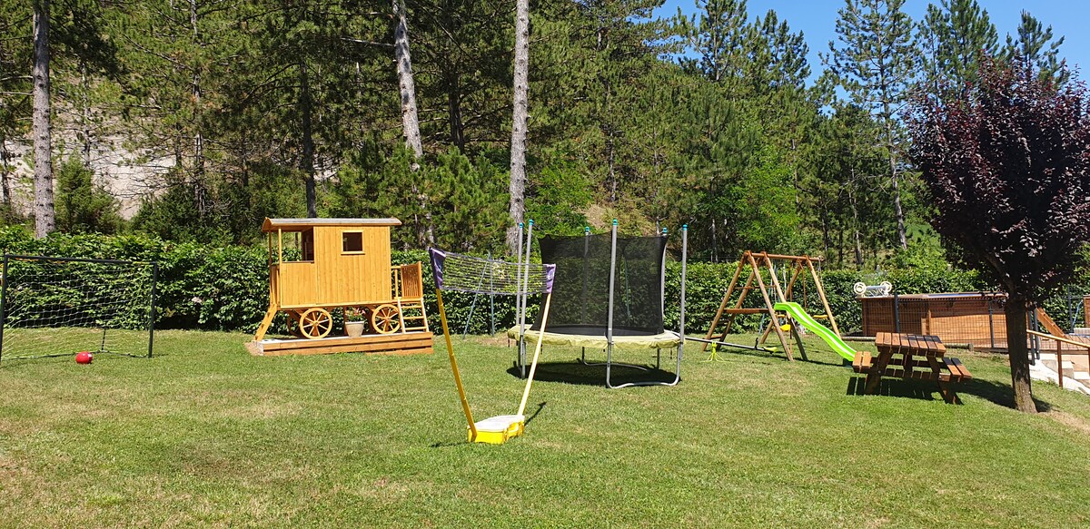 A spacious outdoor area features a wooden playhouse and a trampoline surrounded by lush greenery. A slide and swing set are visible nearby, with a picnic table situated on the grass, providing a cheerful setting for leisure and play.
