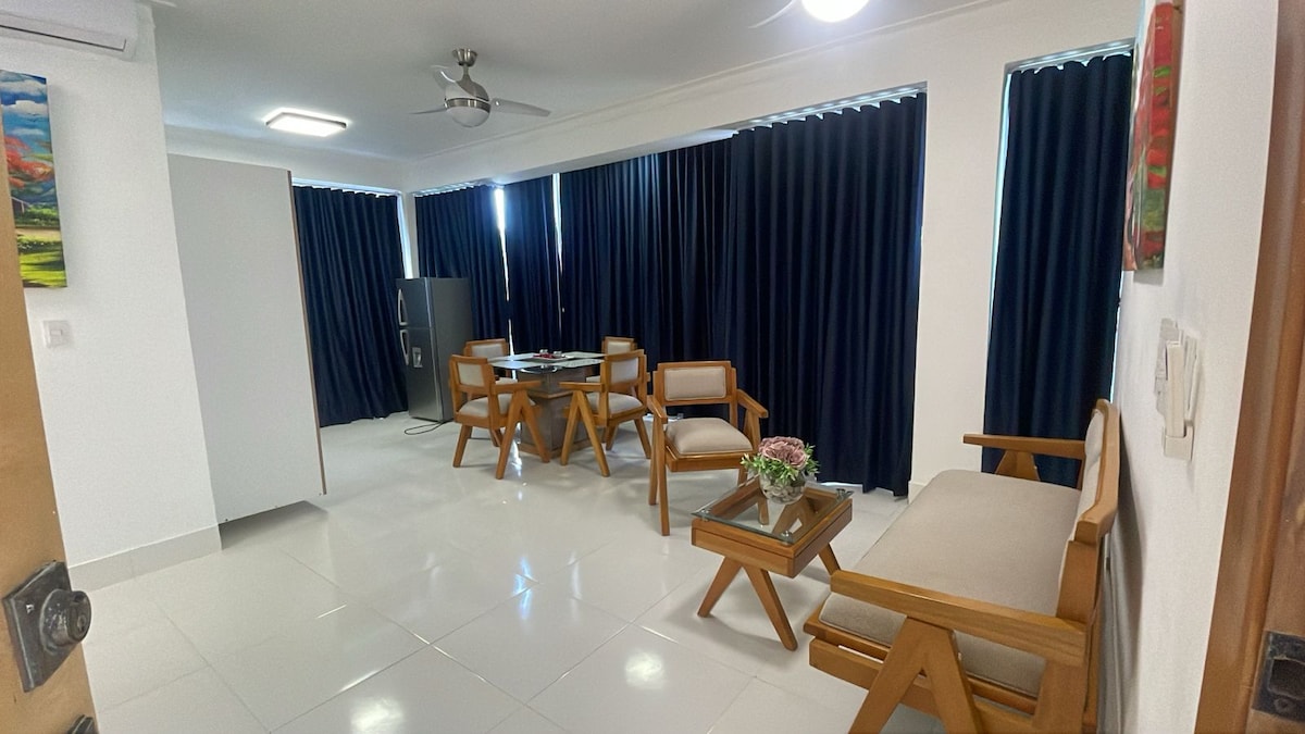 A modern living area features a light-colored tiled floor. Wooden chairs and a table create a cozy seating arrangement, illuminated by natural light from large windows covered with dark curtains. A small decorative vase adds a touch of elegance to the setting.