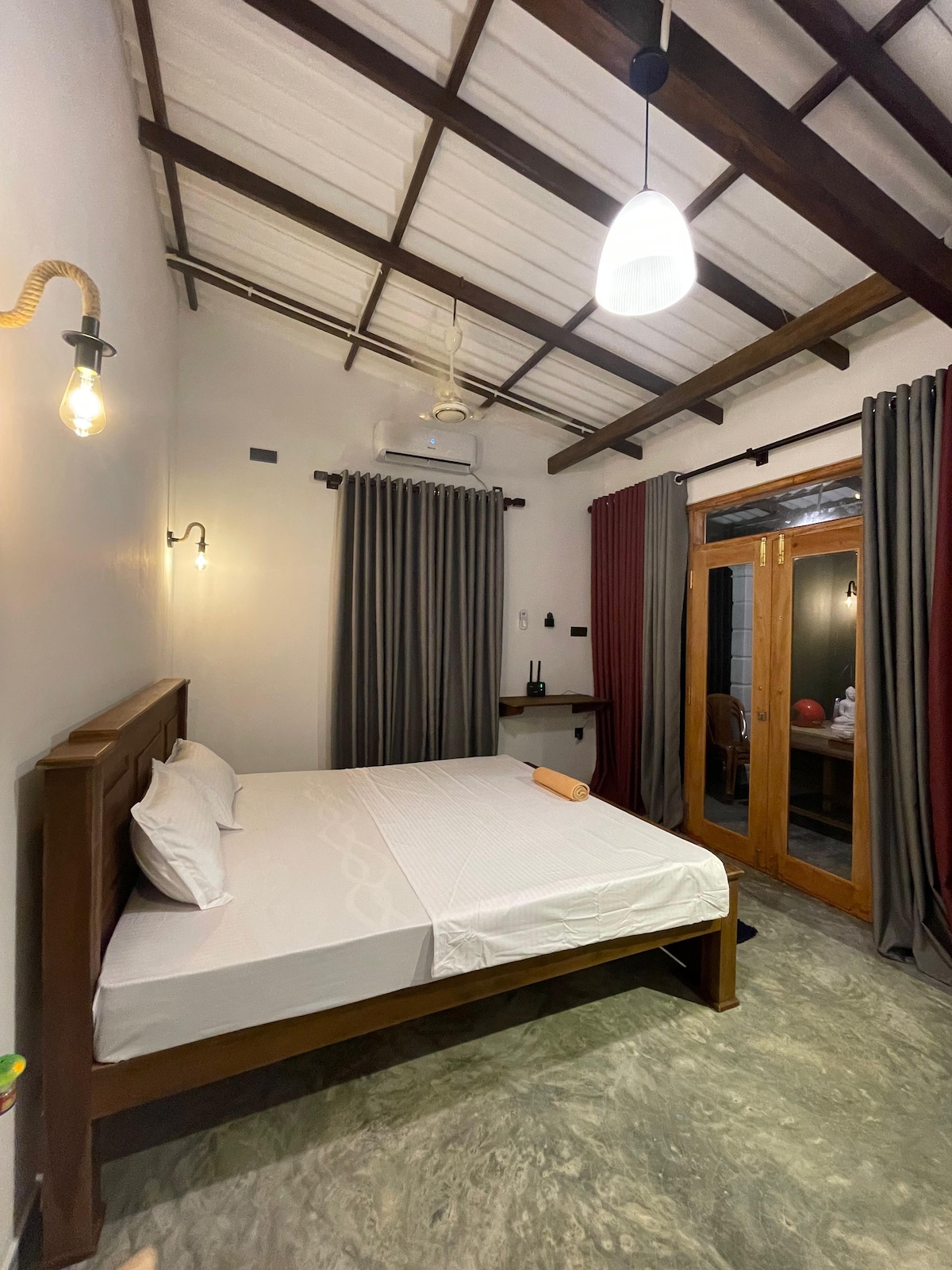 A spacious room features a wooden bed with crisp white linens. Ceiling beams and pendant lights contribute to a cozy ambiance. Dark curtains frame glass doors, leading to an outdoor area, while a small workspace is visible against the wall.