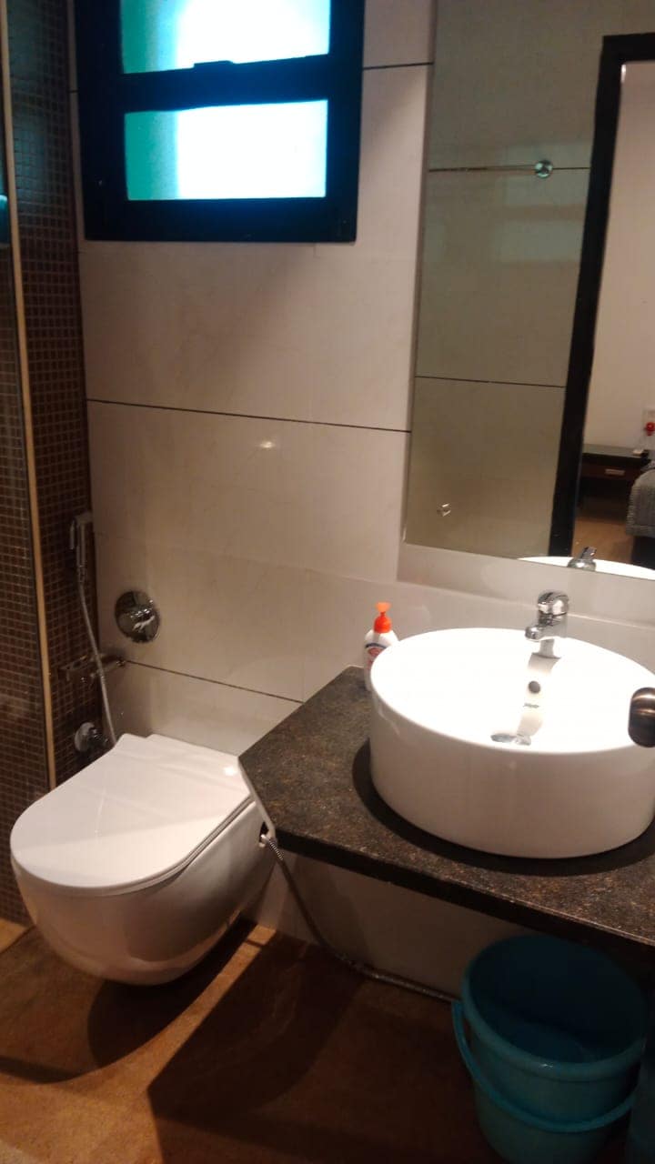 A modern bathroom features a sleek, circular sink atop a dark countertop. A compact toilet is positioned beside the sink, with light reflecting off the glossy wall tiles. A large mirror enhances the sense of space, while a small window allows natural light to enter.