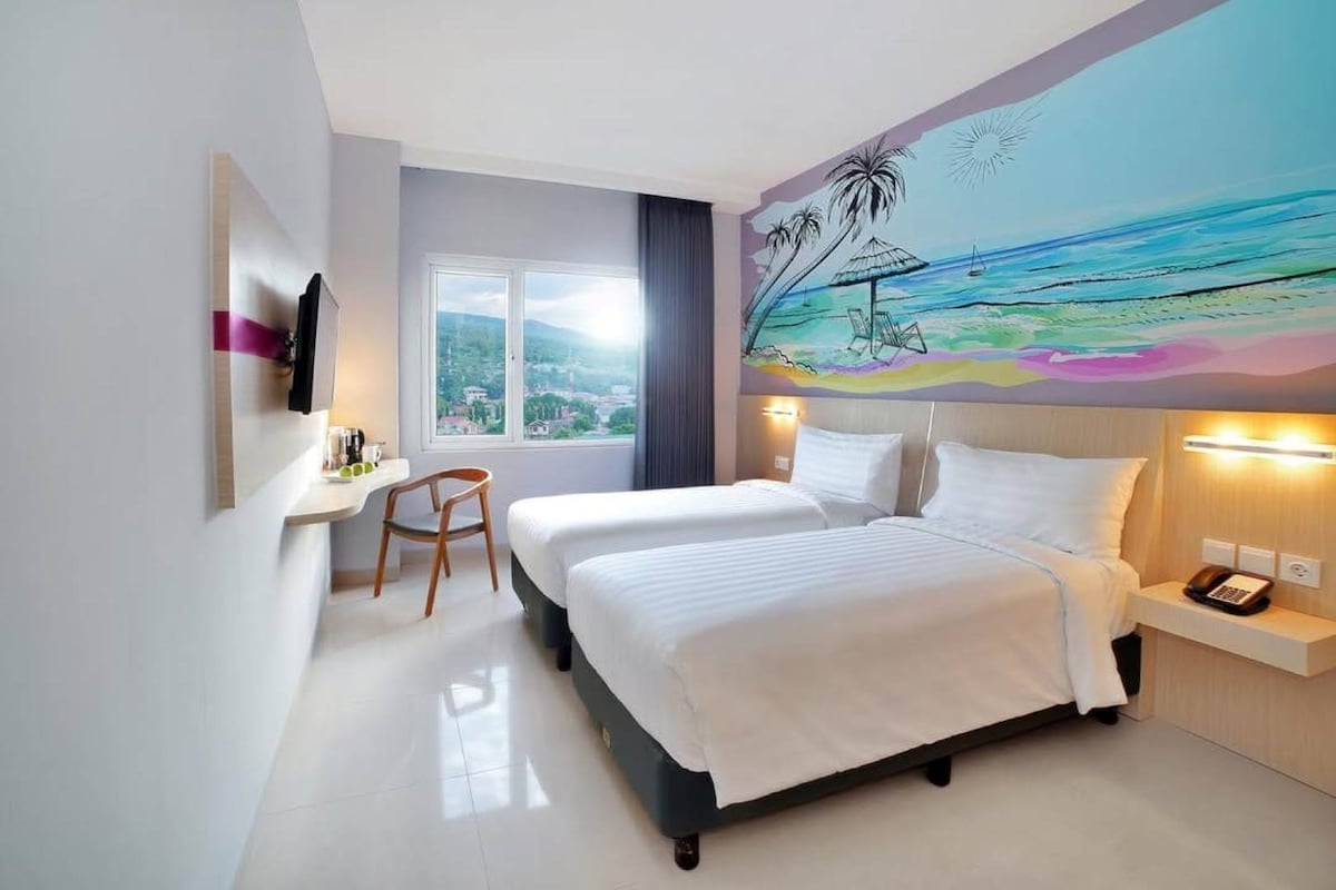 A well-lit guest room features two single beds dressed in crisp white linens. A mural depicting a beach scene adds a pop of color to the wall. A flat-screen TV is mounted opposite the beds, and a small desk and chair are placed near the window.