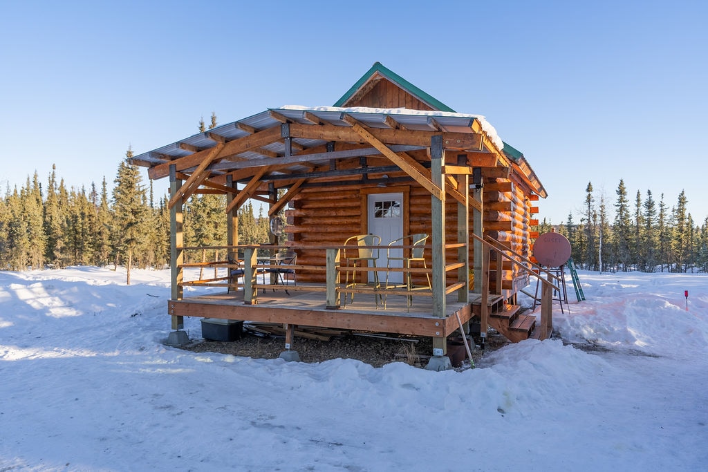 A cozy log cabin features a spacious wooden deck with four chairs and a ramp for easy access. Snow blankets the surrounding area, and tall evergreen trees provide privacy. Bright daylight highlights the rustic charm and warmth of the log construction against the clear winter sky.