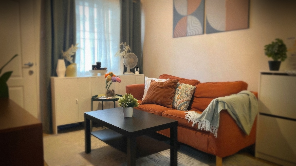 A cozy living area is showcased, featuring an orange sofa adorned with decorative pillows, a light throw, and a coffee table at the center. A potted plant and a floral arrangement are visible on the table. Bright curtains frame the window, complementing wall art with geometric designs.