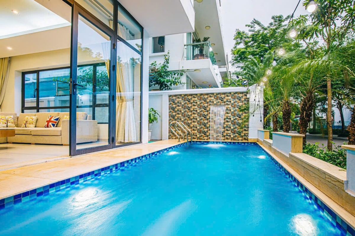 A private pool glistens in a tranquil outdoor area, surrounded by greenery. Large glass doors provide an inviting view of the adjoining lounge space, where comfortable seating is arranged. Soft lighting enhances the serene atmosphere in the evening setting.