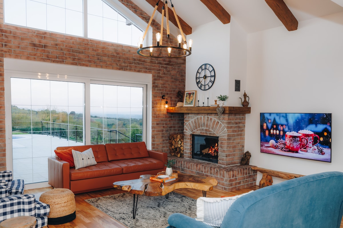 A cozy living area features a mix of seating options, including a spacious orange couch and a blue armchair. A rustic coffee table rests on a patterned rug, while a large flat-screen TV is mounted on the wall. Natural light floods in through expansive windows.