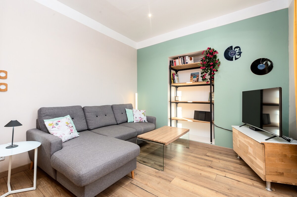 A spacious living area features a contemporary grey sectional sofa adorned with floral cushions. A glass coffee table sits in the center, complemented by a minimalist lamp. A shelf with books and decorative items is situated against a green accent wall, while a flat-screen television is positioned on a light wood console.