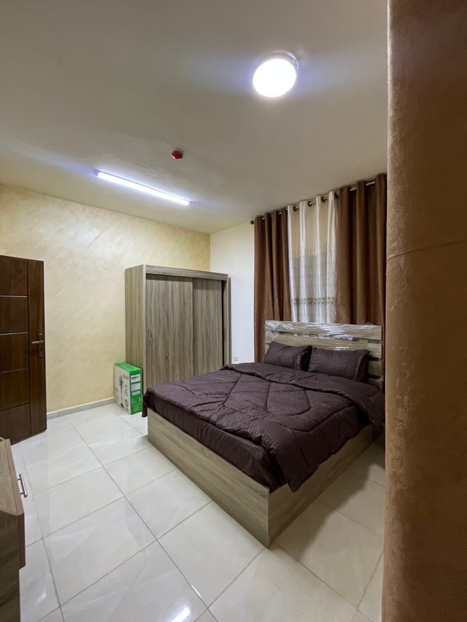 A well-appointed bedroom features a spacious bed dressed in dark bedding. A large wardrobe stands against the wall, while curtains drape over the window. Soft lighting enhances the room's ambiance, and tiled flooring adds a modern touch.